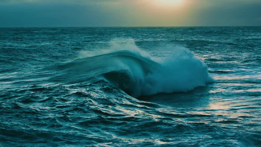 A majestic ocean wave builds and crashes in dramatic slow motion as the sun sets on the horizon