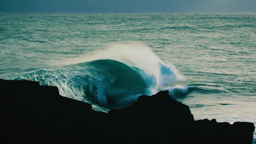 A powerful ocean wave builds and crashes in dramatic super slow motion, framed by a dark, silhouetted rocky shoreline