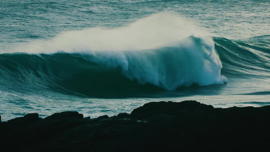 A powerful ocean wave builds and crashes in dramatic slow motion behind a rocky shoreline