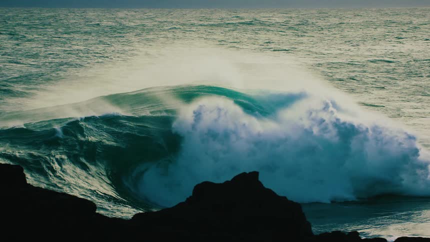 A massive teal ocean wave curls and crashes in dramatic slow motion with silhouetted rocks in the foreground