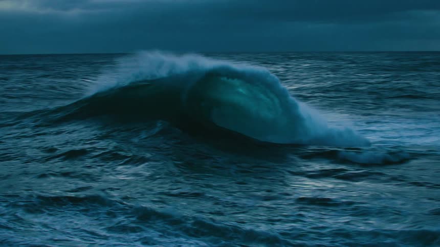 A powerful ocean wave swells and crashes in dramatic slow motion under a dark and moody sky