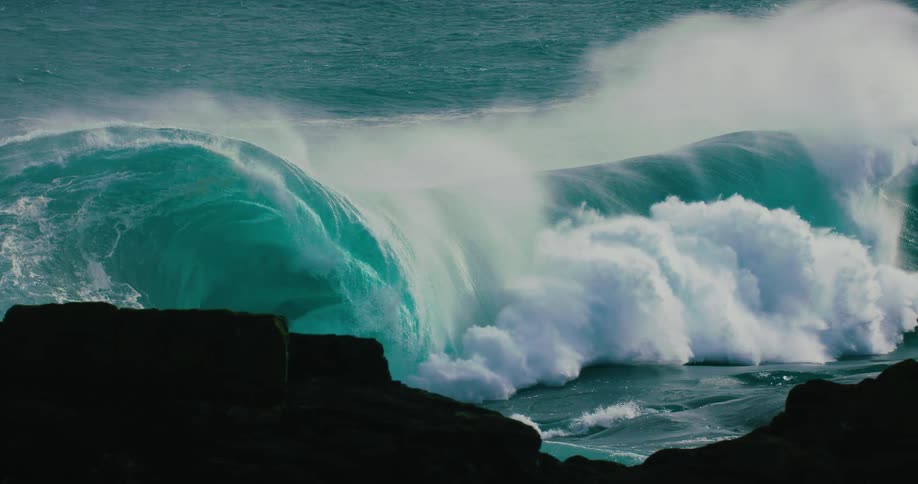 A powerful turquoise ocean wave builds and crashes in dramatic slow motion behind a silhouetted rocky coastline