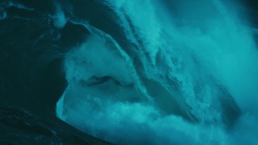 A massive, powerful teal-colored ocean wave builds and crashes in dramatic slow motion