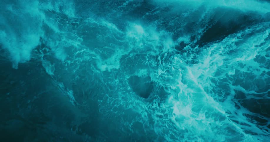 A dramatic slow-motion close-up captures a massive, turquoise ocean wave as it builds and crashes with immense power