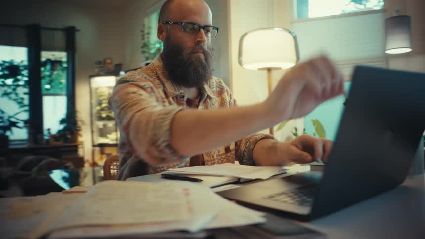 A man with a beard and glasses opens his laptop to work from his cozy home office