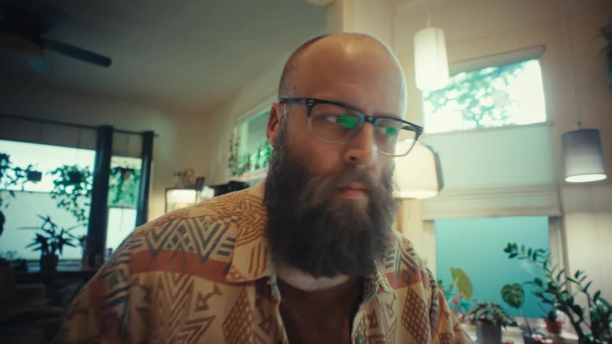 A slow push-in shot focuses on a bearded man with glasses as he works diligently on his laptop from his cozy, plant-filled home