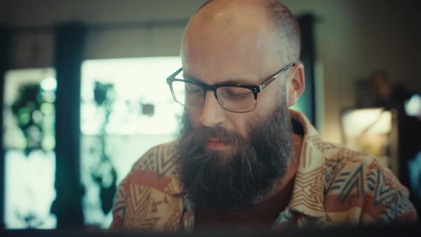 A cinematic zoom-out shot reveals a bearded man with glasses working intently on his laptop in his home office