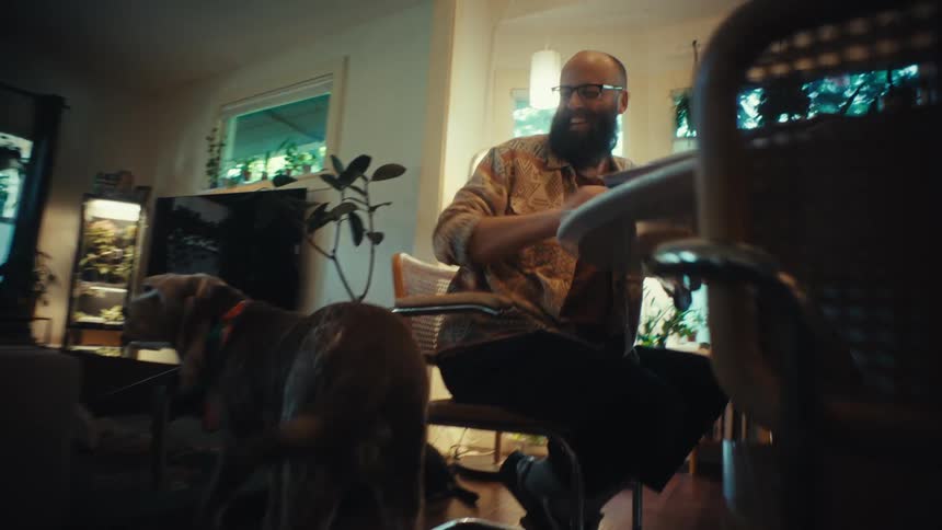 A dog walks over to its owner, a bearded man happily working on his laptop at a table in his cozy home