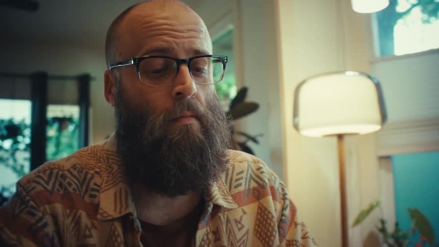 Low-angle shot reveals a focused, bearded man with glasses working on his computer in his living room