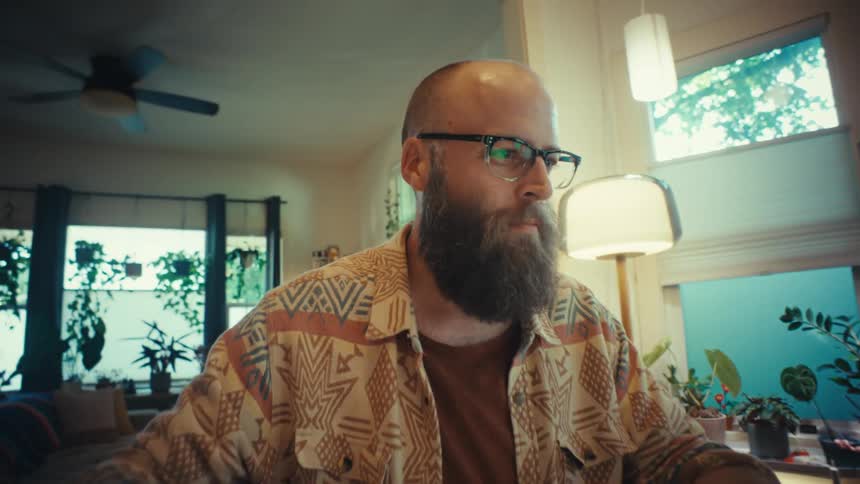 A focused, bearded man wearing glasses works from home on his laptop in a cozy, plant-filled office