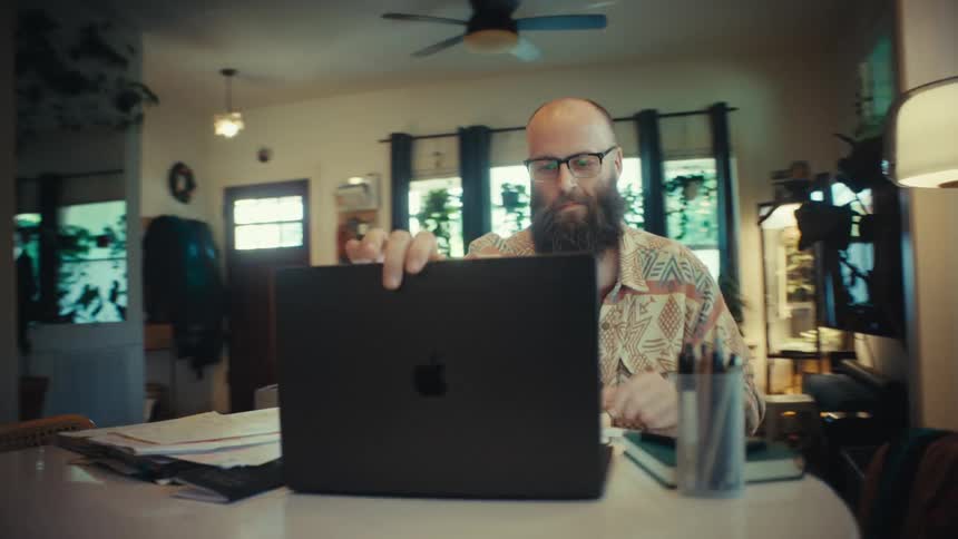 A content man with a beard closes his laptop, finishing his work day from his plant-filled home office
