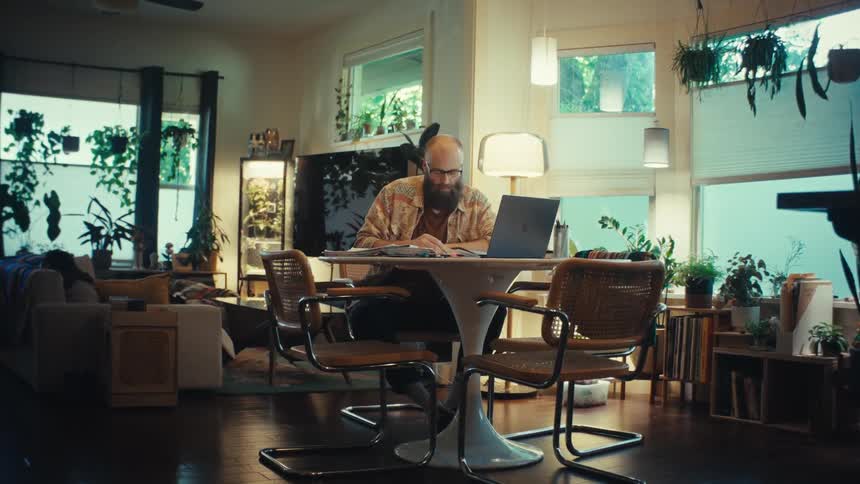 A man with a beard works on his laptop in a cozy, plant-filled living room as the camera slowly pushes in