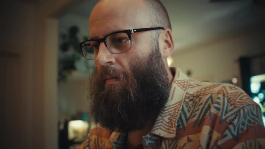 A focused bearded man wearing glasses works on his laptop computer at home, the camera slowly pushing in on his face