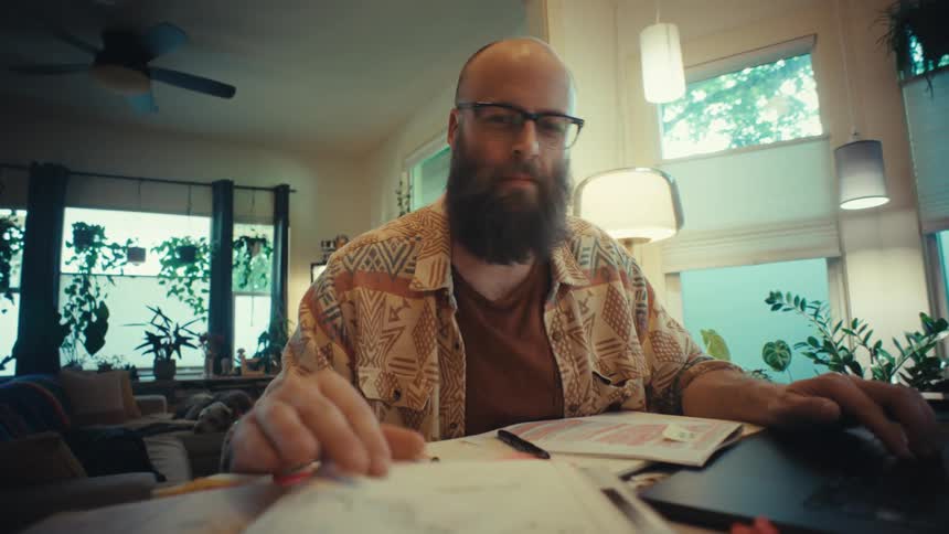 A bearded man wearing glasses works on his laptop and flips through a book while sitting at a table in his cozy, plant-filled home