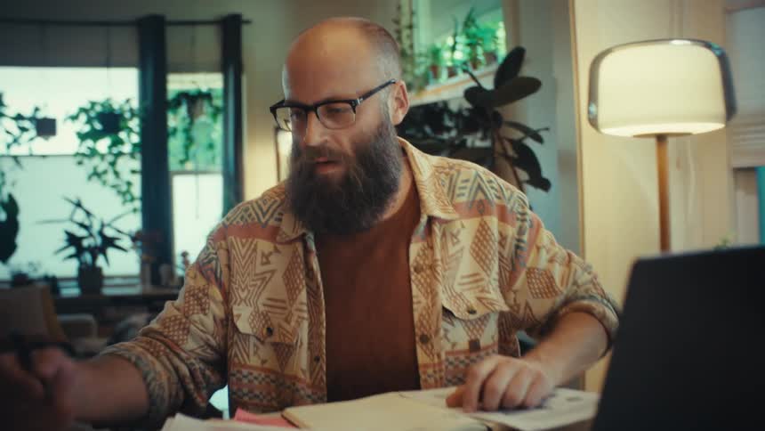 A focused bearded man meticulously sorts through paperwork at his home desk before turning his attention to his laptop