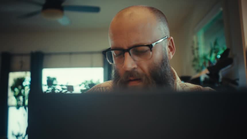 A cinematic close-up of a focused man with a beard and glasses working on his laptop in a home office