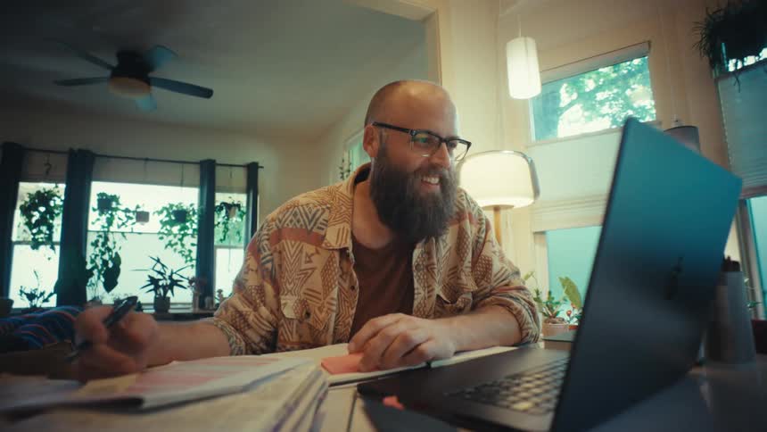 A bearded man wearing glasses smiles while working from home, taking notes from his laptop at a table in his sunlit, plant-filled house