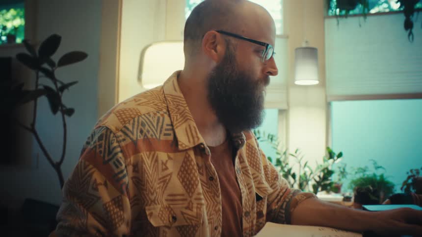 A bearded man with glasses works from home on his laptop, focusing intently on his task as the camera slowly zooms in