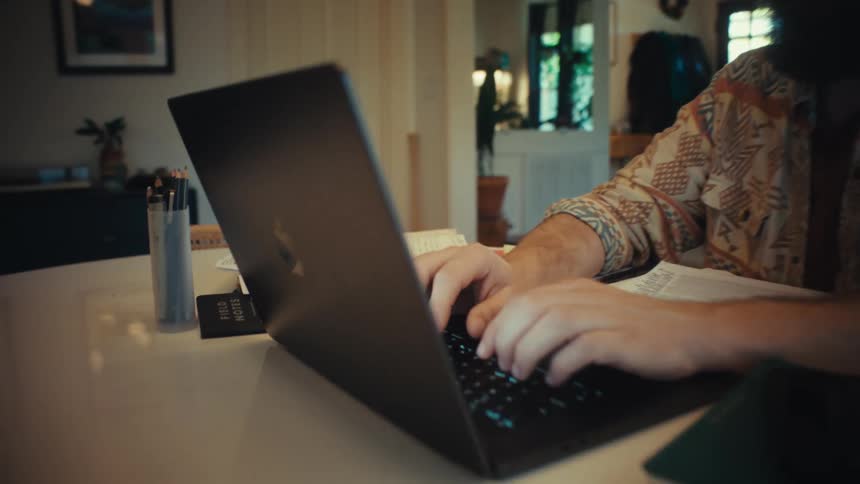 A slow push-in shot shows a focused, bearded man working from home on his laptop at the dining table, ending on a close-up of his hands typing