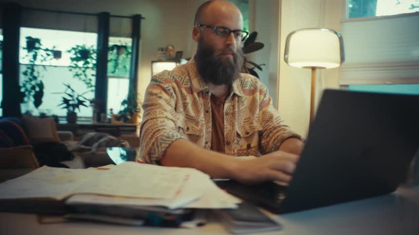 A bearded man in glasses opens his laptop at a dining table and begins to type, working from his cozy, plant-filled home