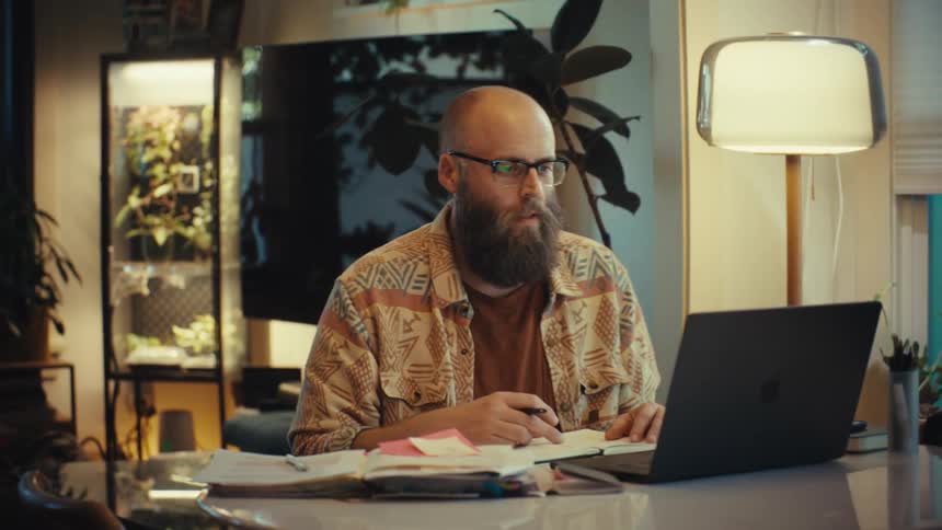 A bearded man wearing glasses works on his laptop in a cozy home office in the evening, taking notes and participating in a video call