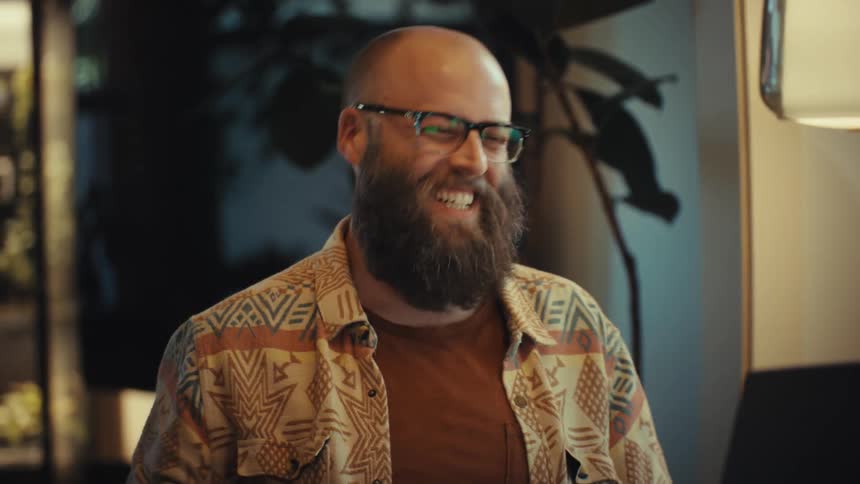 A happy bearded man with glasses laughs enthusiastically during a video call on his laptop in a cozy room