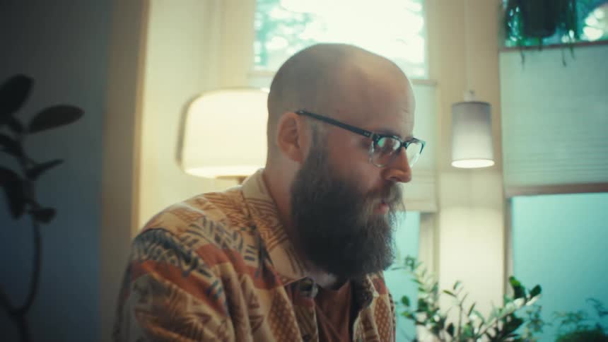 Slow push-in shot of a bearded man with glasses studying intently at his laptop in a cozy, plant-filled apartment