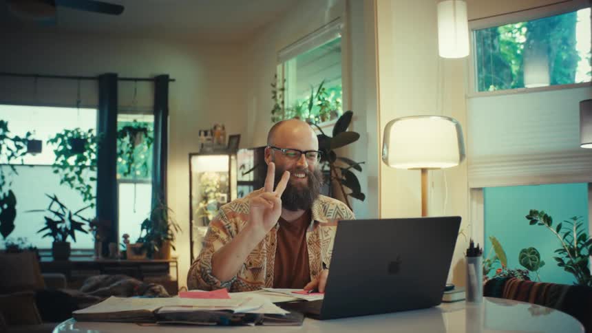 A friendly bearded man works from home, smiling and giving a peace sign during a video call in his cozy, plant-filled living room