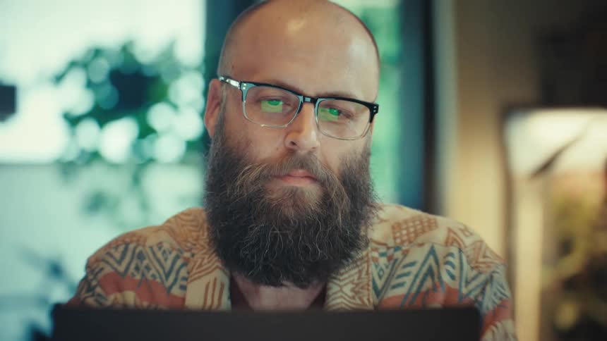 A stylish bearded man in glasses concentrates while working on his laptop in a plant-filled room