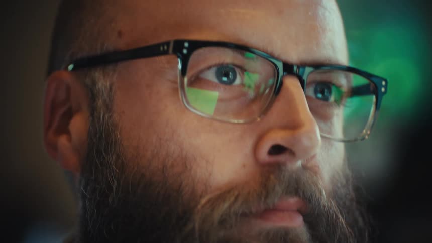 Intense close-up of a focused bearded man in glasses with a screen's reflection in his eyes