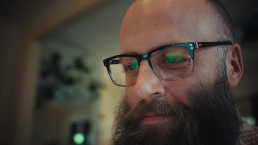 A happy bearded man with glasses smiles while working on his laptop, with the camera slowly zooming in on his face