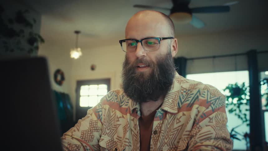 Close up of a smiling bearded man in glasses working remotely on a laptop computer