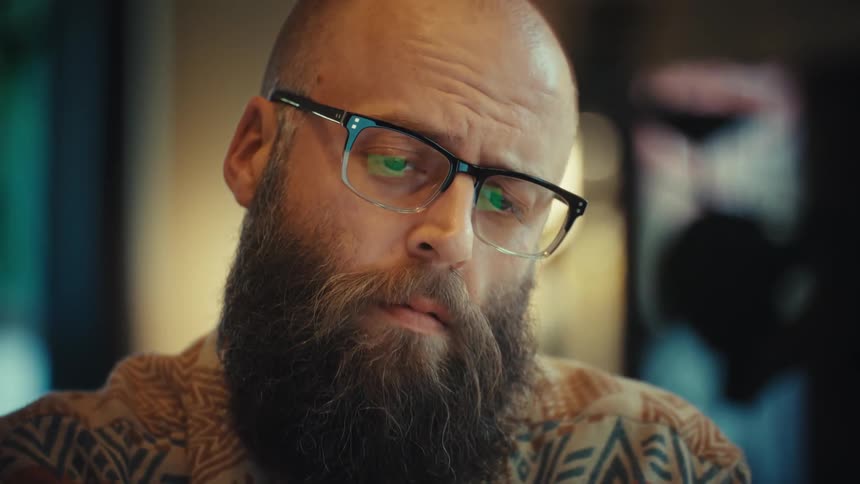 A focused bearded man wearing glasses works on his laptop in a home office as the camera slowly zooms into a close-up