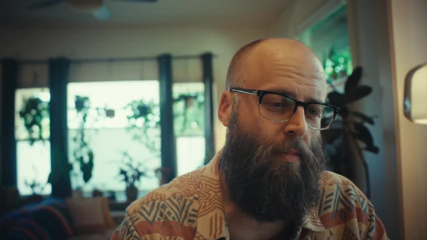 Close up of a focused, bearded man with glasses working on his laptop in a home office before closing it
