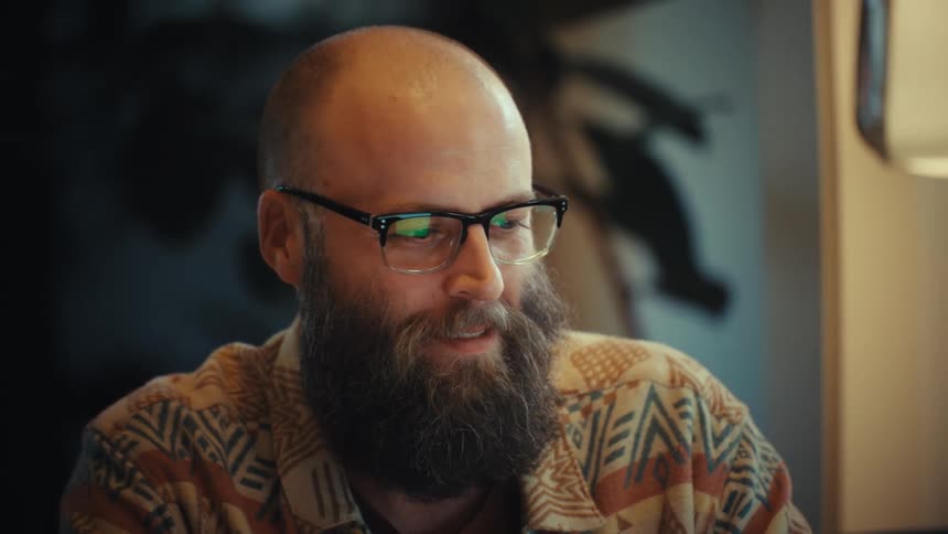 A happy bearded man with glasses smiles and laughs while on a video call on his laptop from his cozy home office