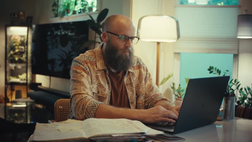 A slow zoom-in on a focused bearded man in glasses typing on his laptop while working from his cozy home