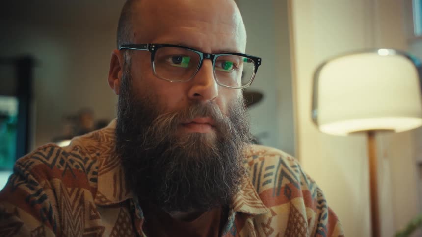 A bearded man with glasses focuses intently on his laptop, the camera slowly zooming in on his concentrated face in a warm home office