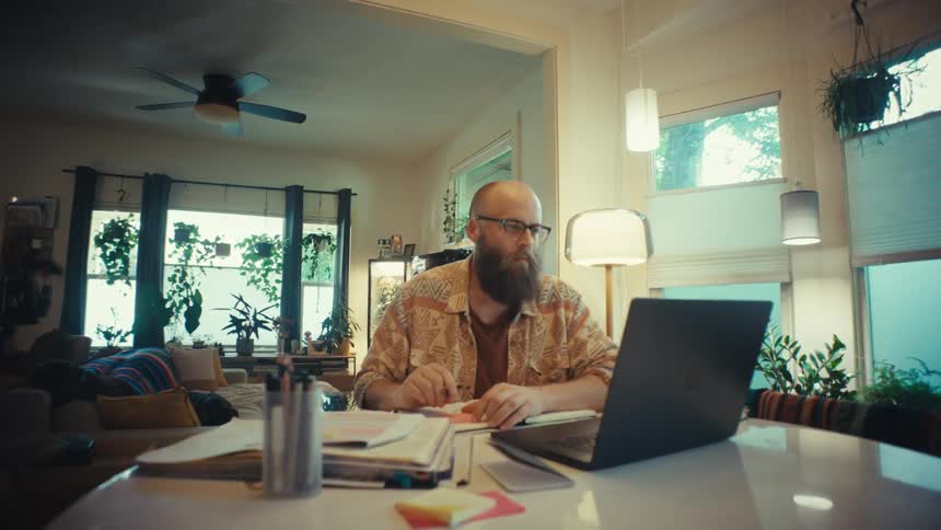 A man with a beard and glasses works remotely from his plant-filled bohemian home, taking notes and then concentrating on his laptop