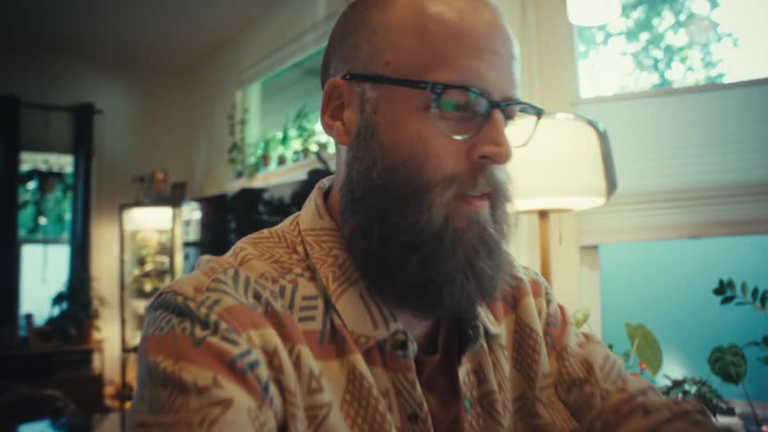 A bearded man in a patterned shirt and glasses opens his laptop at a table in his cozy, plant-filled home to begin work
