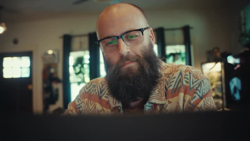 A focused, bearded man wearing glasses smiles while working from home on his computer as the camera slowly pulls away