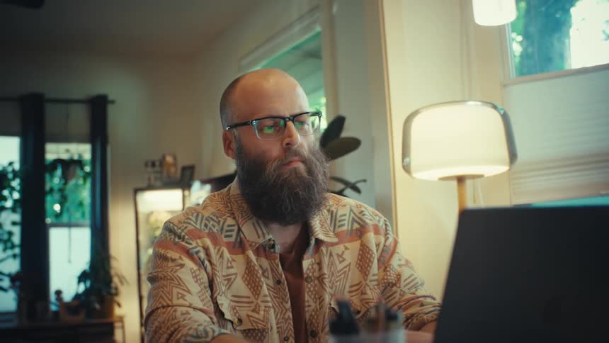 A serious bearded man with glasses works intently on his laptop in his home office as the camera pushes in for a close-up