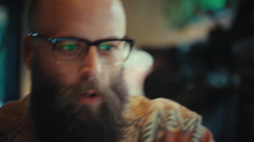 An enthusiastic bearded man wearing glasses gets excited and claps while working on his laptop from home
