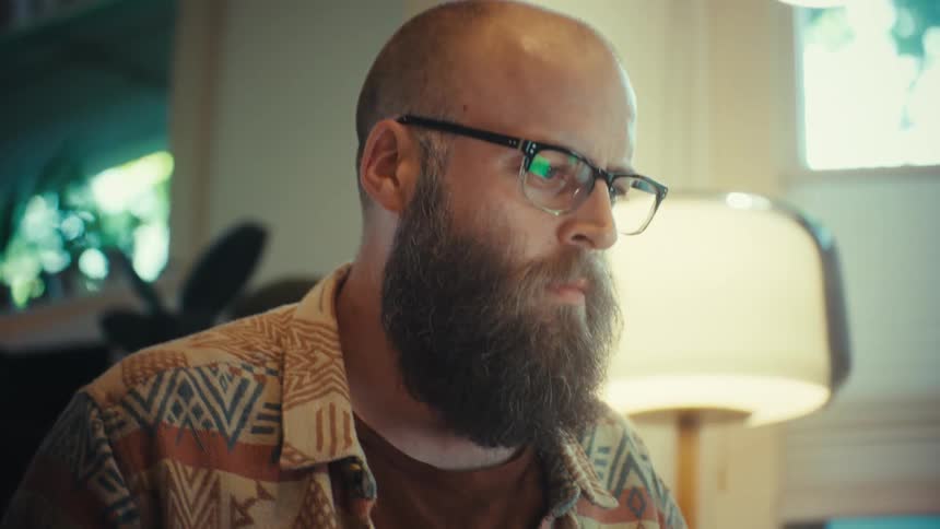 A serious bearded man with glasses concentrates while reading important documents in his home office