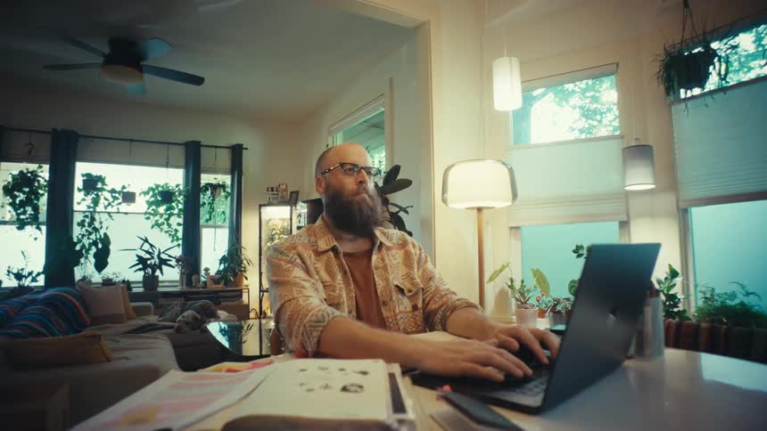 A focused bearded man works from his stylish, modern home on a laptop, with the camera slowly pushing in to a thoughtful close-up