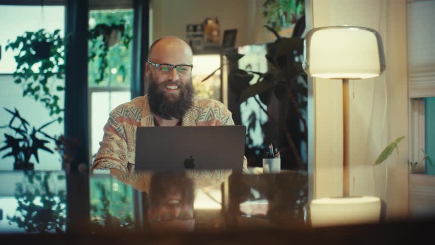 A smiling bearded man in glasses enjoys working from home on his laptop in a cozy, plant-filled room