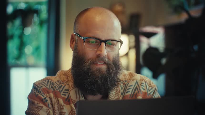 A focused bearded man wearing glasses works intently on his laptop in his home office as the camera slowly zooms in