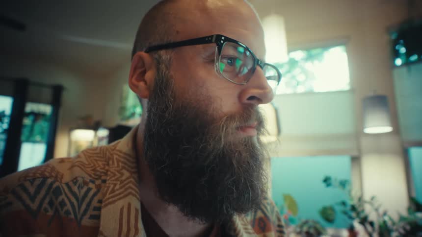 A close-up camera move reveals a bearded man with glasses working diligently on his laptop in a cozy home office