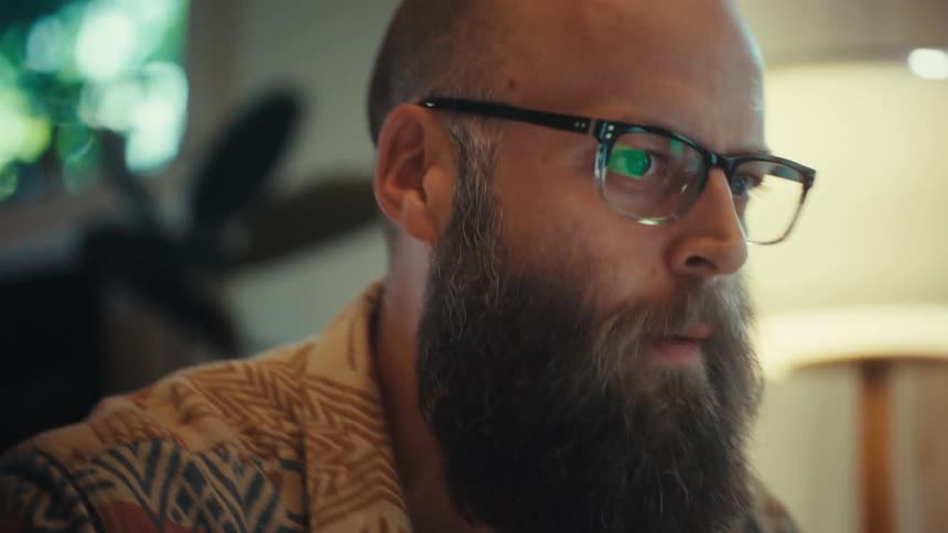 An intense bearded man wearing glasses argues passionately during a video call on his laptop in a home office