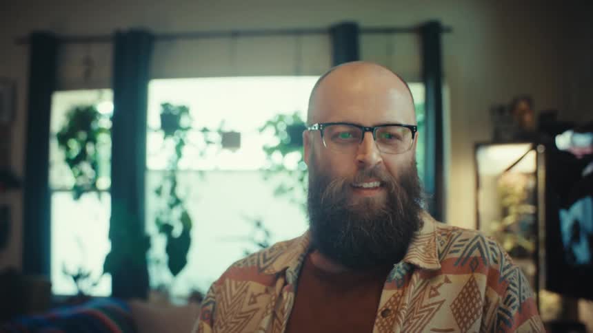 A stylish bearded man in glasses works from home on his laptop, smiling as the camera slowly pushes in for a close-up