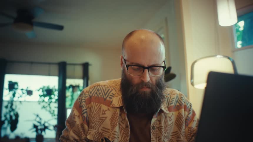 A close up reveals a focused, bearded man with glasses working on a laptop at his home desk